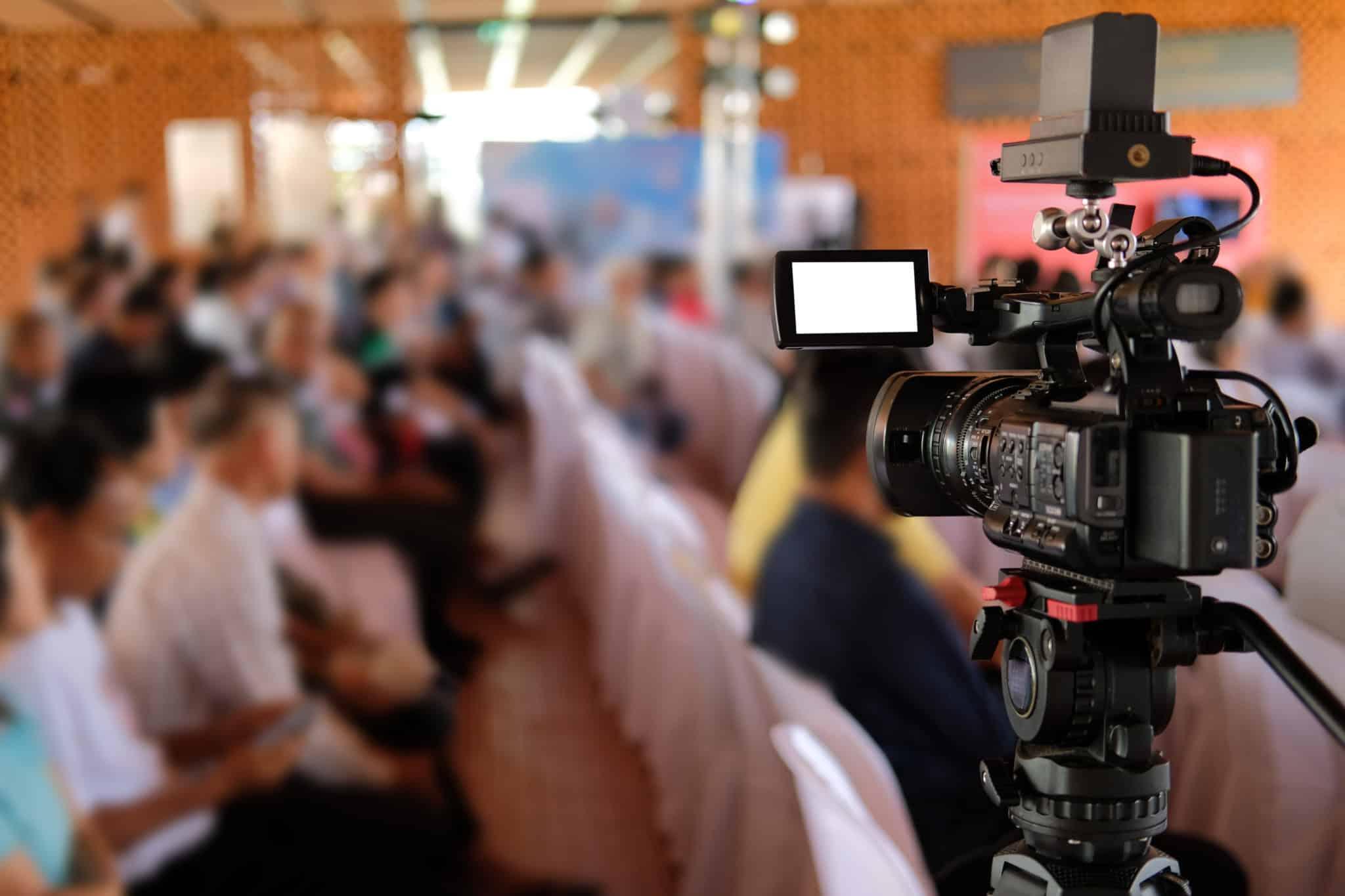 An image of a camera recording in the right foreground of the image with a group of people gathered in a hall in the background for customer spotlight blog.
