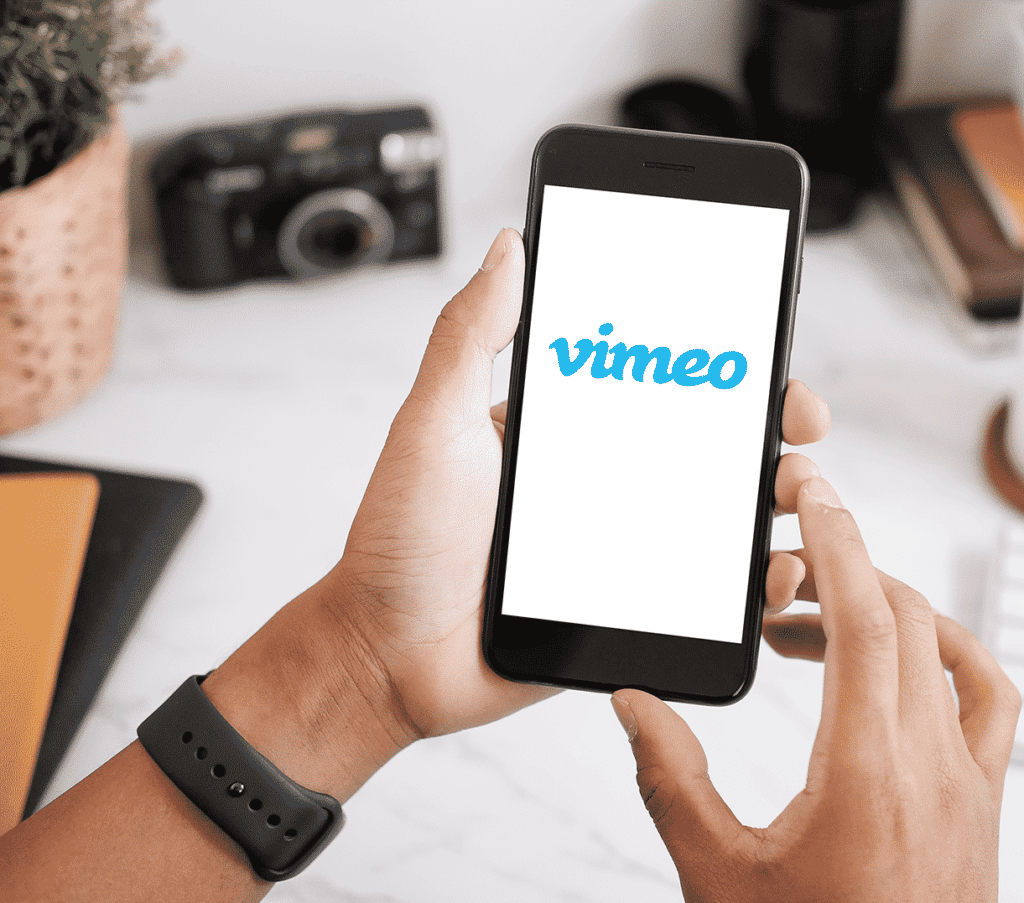 two hands holding up a smartphone with the vimeo app loading with a desk and camera in the background