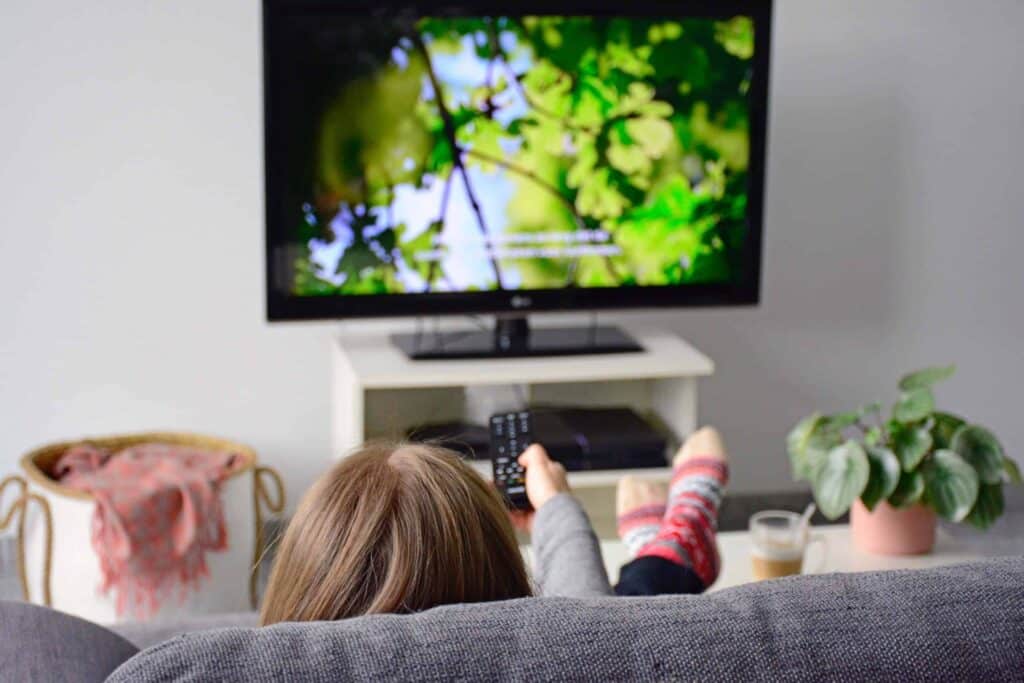 A girl sitting on the couch with tv remote pointed at the tv screen that has picture of tree branches and open captions
