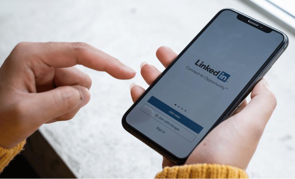 a pale hand holds a smartphone that shows the LinkedIn application loading, the logo reading LinkedIn with the word 