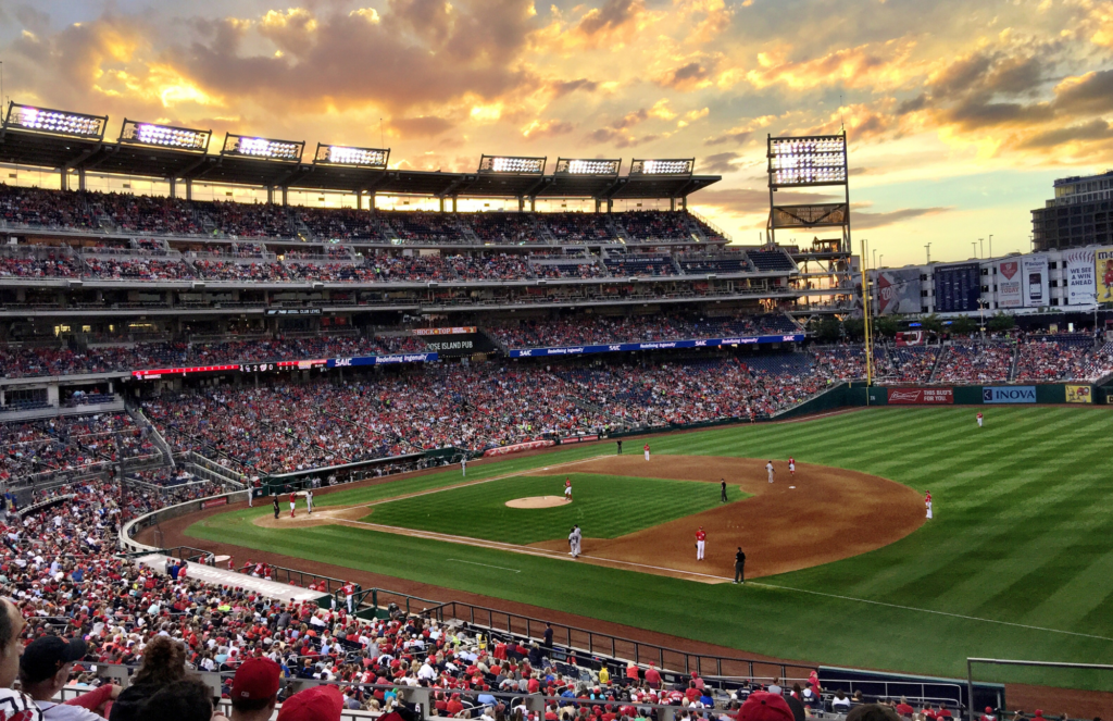 View of a packed baseball stadium from far off seats on the first base side of the field. Players are on the field, the sky is cloudy with a sunset.
