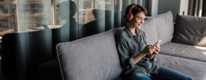 Image of nice young smiling woman using smartphone and headphones while sitting on couch at living room