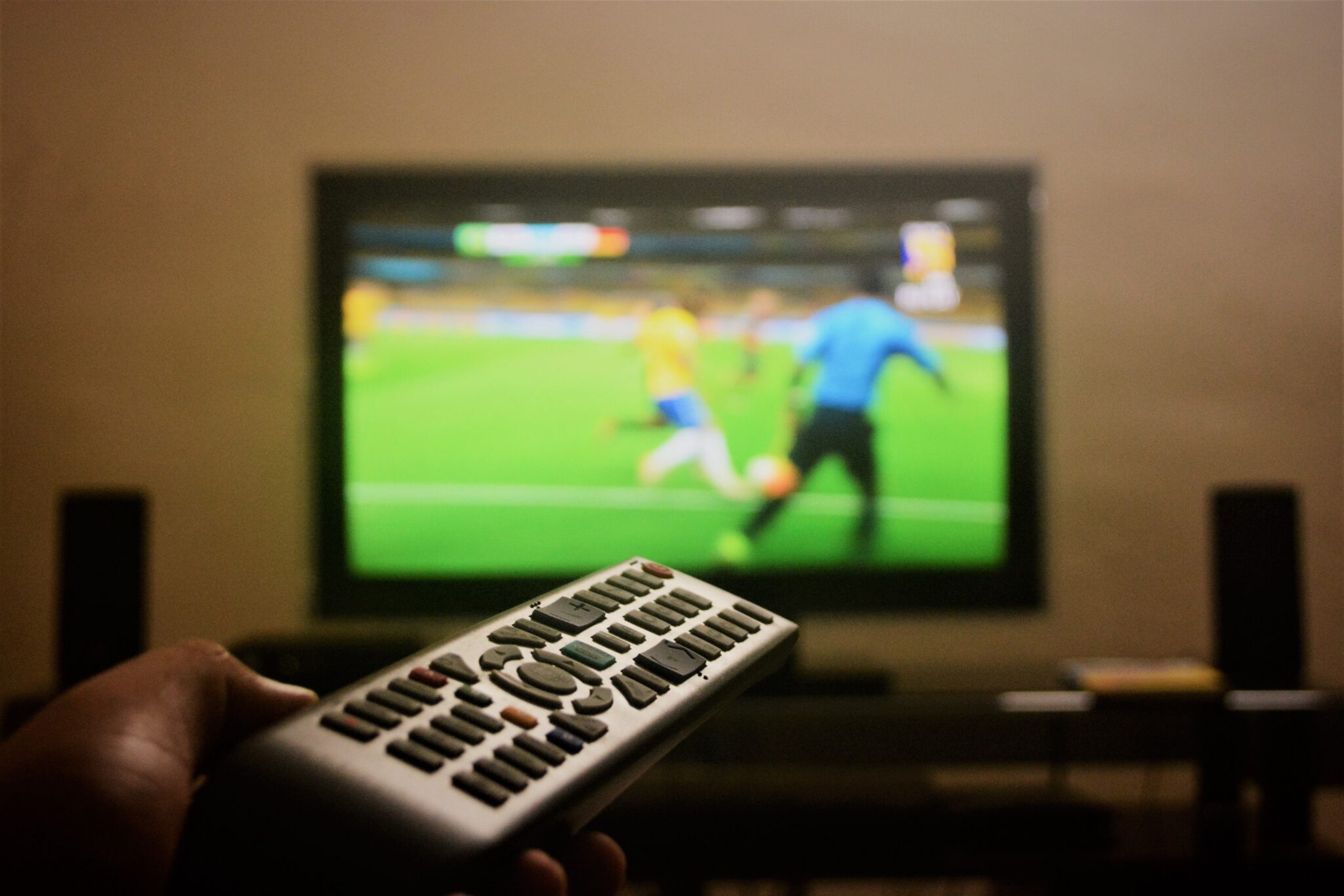 Close up on television remote control with a blurred television set in the background. A soccer game can be seen on the set.