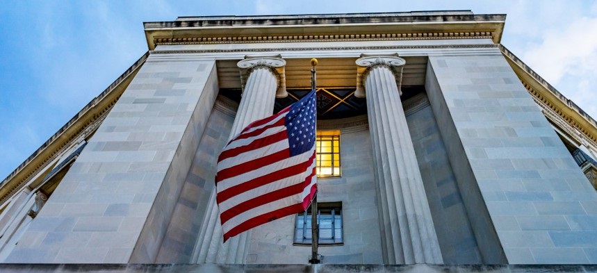 An exterior photo of the DOJ with an american flag flying in front of two tall white columns.