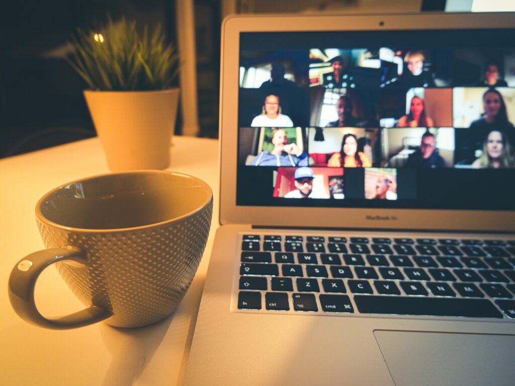 A cup of coffee sits next to an open laptop on a desk. The laptop is viewing a conference call with multiple people on the screen for the FCC blog