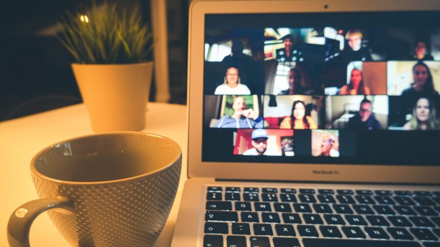 A cup of coffee sits next to an open laptop on a desk. The laptop is viewing a conference call with multiple people on the screen for the FCC blog