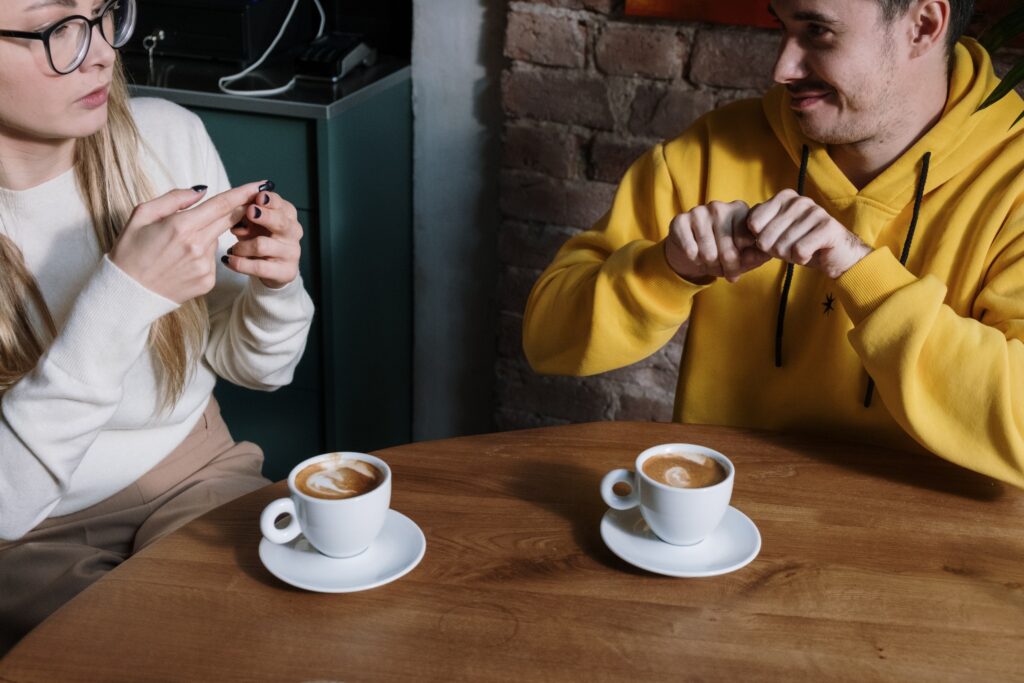 A man and a women seated at a table with coffee cups converse using sign language.