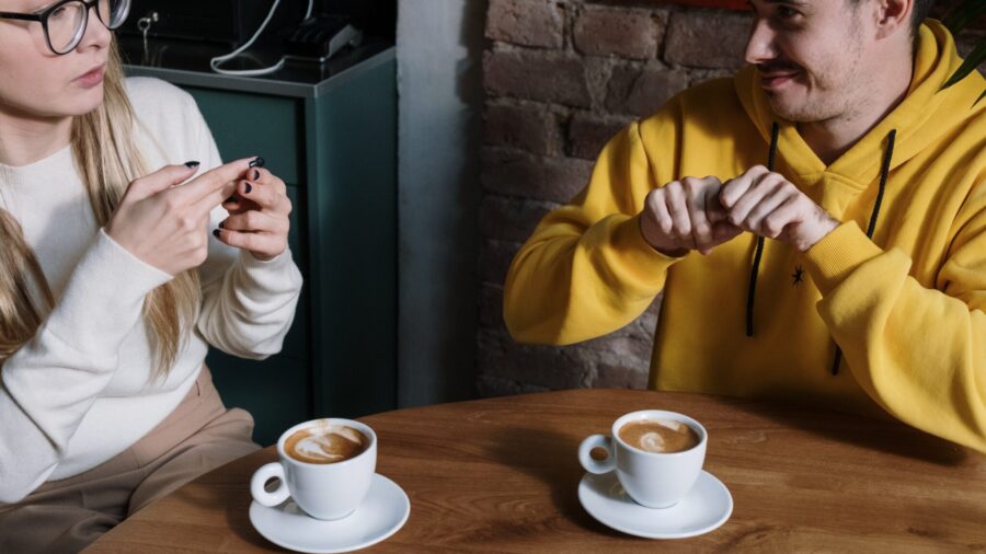 A man and a women seated at a table with coffee cups converse using sign language.