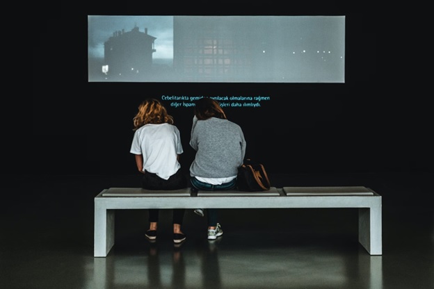Two people sitting on a bench, seen from behind, watching a movie screen with captions running on the bottom of the screen.