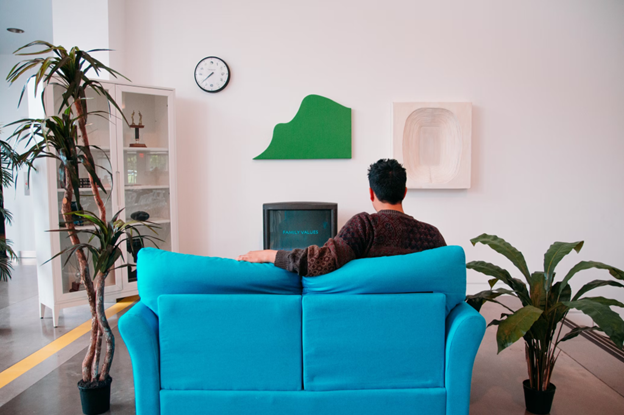 A man sit on a bright blue couch, his back toward the viewer, and watches television