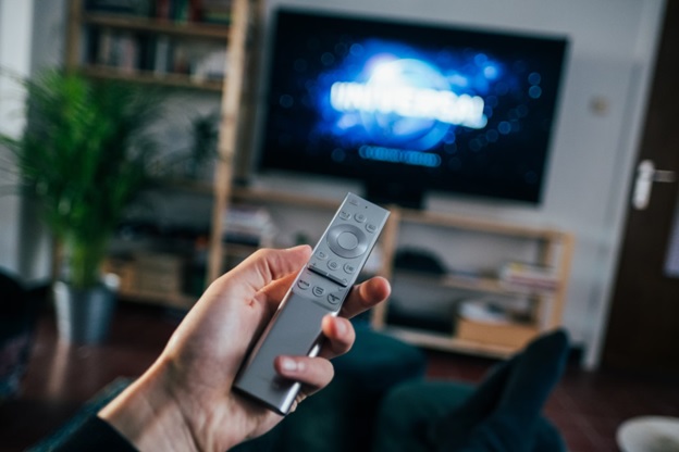 Close up of a hand holding a television remote control. In the background is a blurred television screen