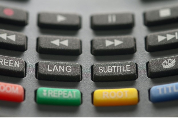 Close up of a television remote control, showing numerous and multicolored buttons. Prominently displayed in the center is a button with the word 'subtitle.'