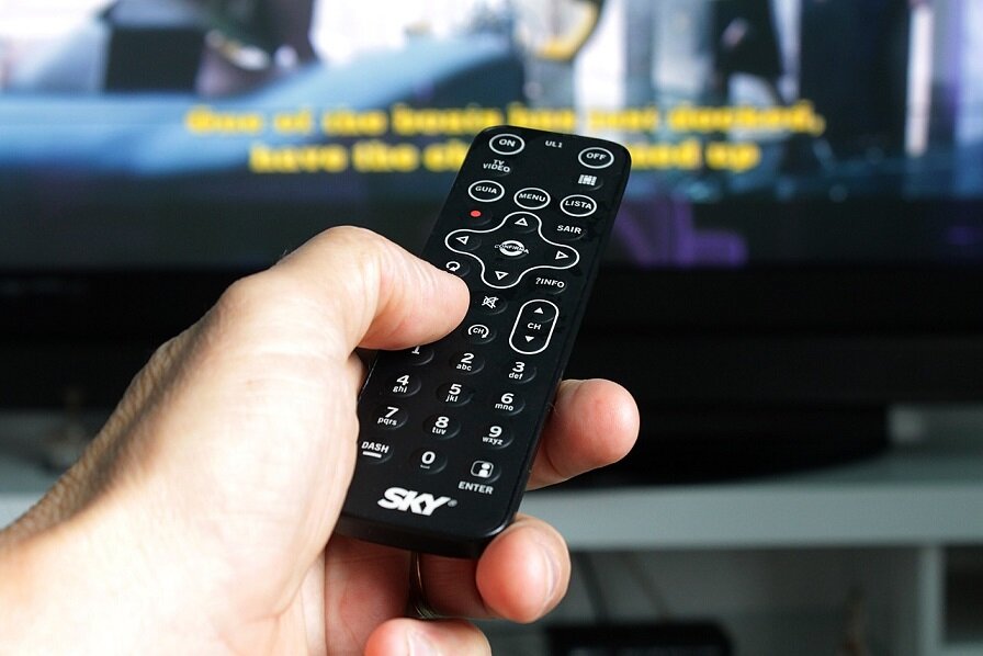 A hand holding a remote control, pointing it at the television. On a blurred screen you can see yellow captions running across the bottom of the image