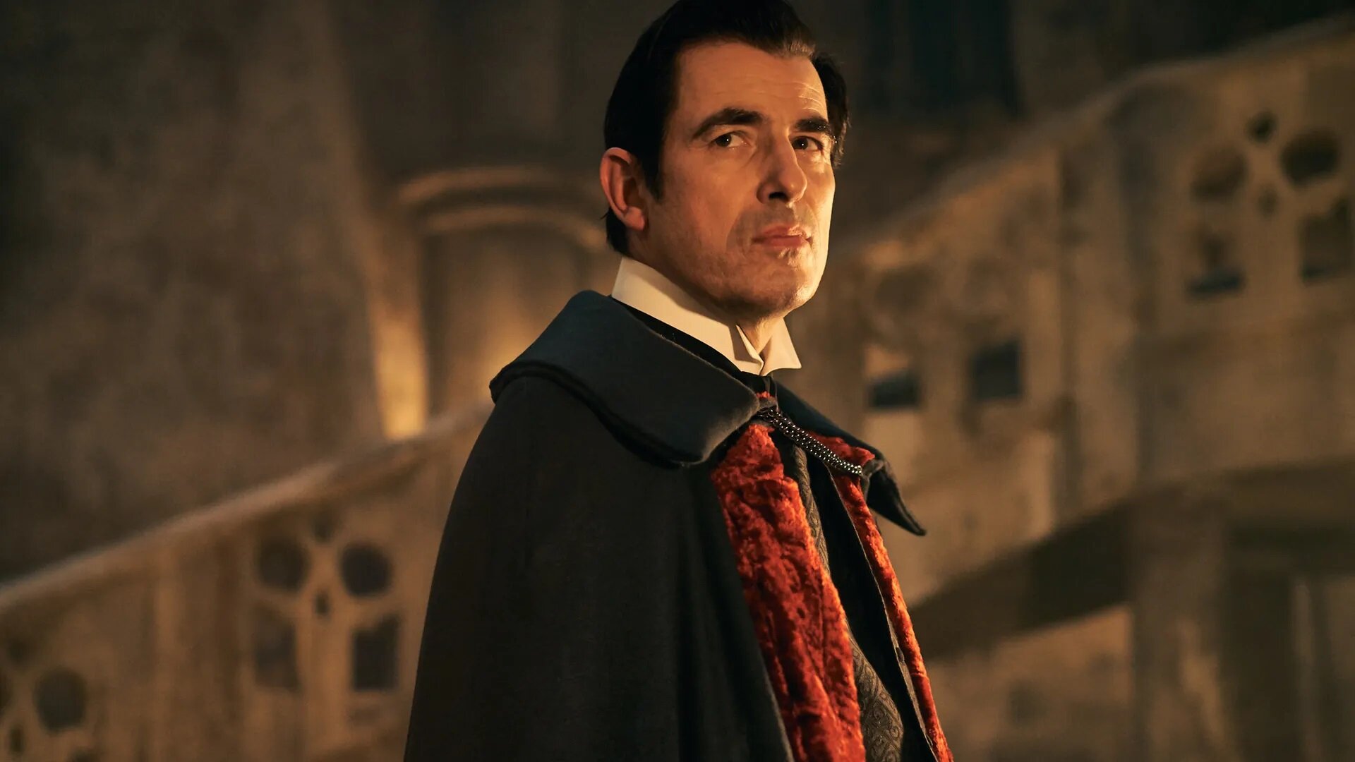 Dracula, dressed in a black cloak with red scarf looks towards the camera