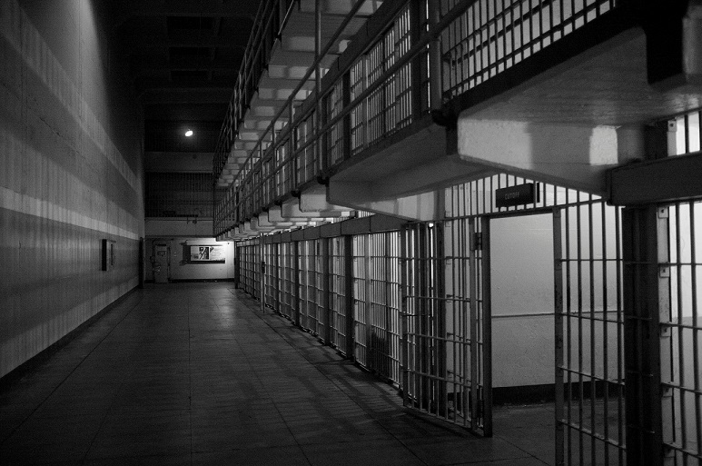 Black and white image looking down a prison corridor with jail cells along the right side of the hallway. For ADA blog