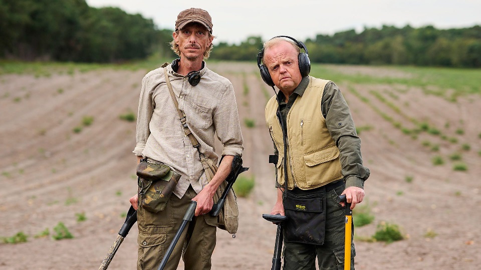 Two men stand in a field and stare directly at the camera. They are each holding metal detectors. For the TV show 'Detectorists'