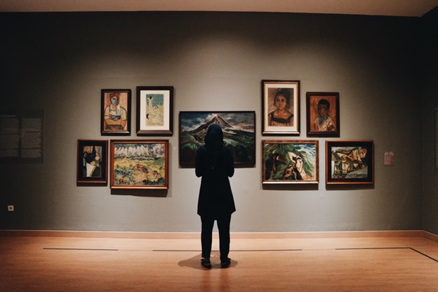 A person stands with their back towards the camera and faces a series of pictures on a gallery wall