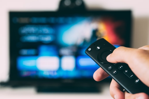 Hand holding a remote control, pointing it towards a blurred television screen in the background