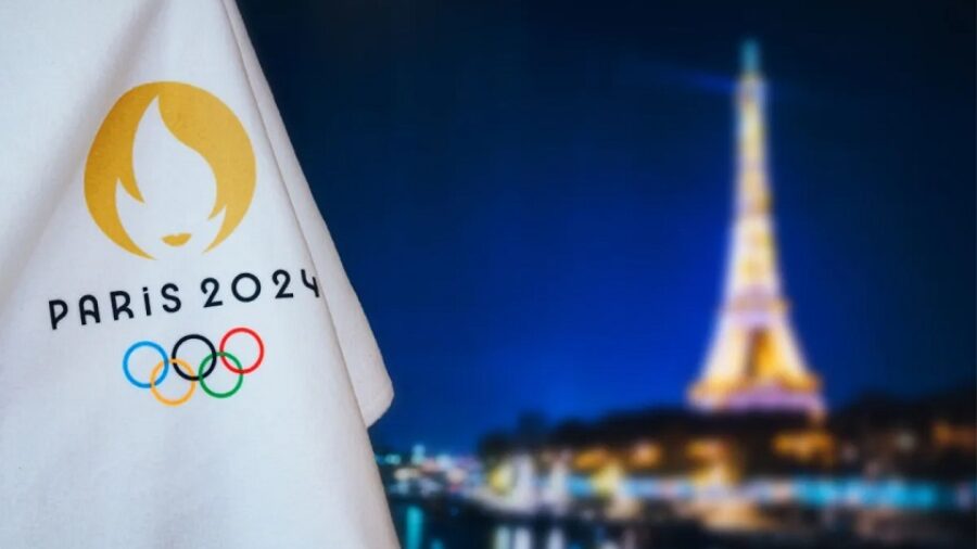 An Olympic flag in the foreground reads Paris 2024 and includes the Olympic rings. Blurred in the background is the Eiffel Tower at night