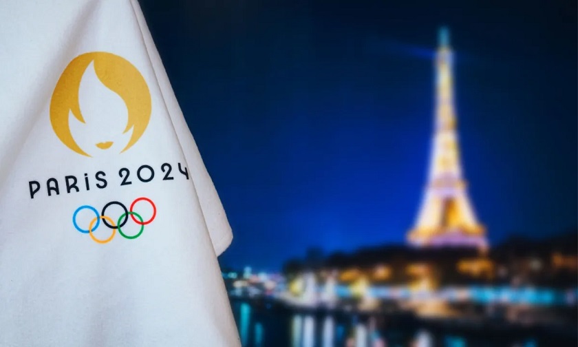 An Olympic flag in the foreground reads Paris 2024 and includes the Olympic rings. Blurred in the background is the Eiffel Tower at night