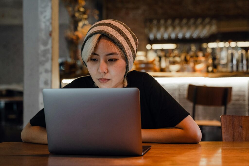 A woman in a knit wool cap sits at restaurant tables and looks at her laptop.